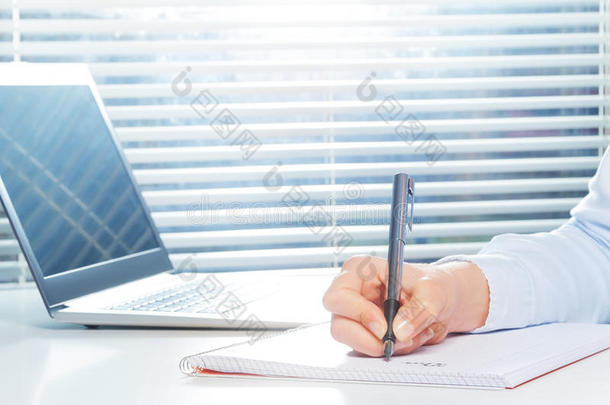 手在笔记本上写字，笔在笔记本电脑旁边