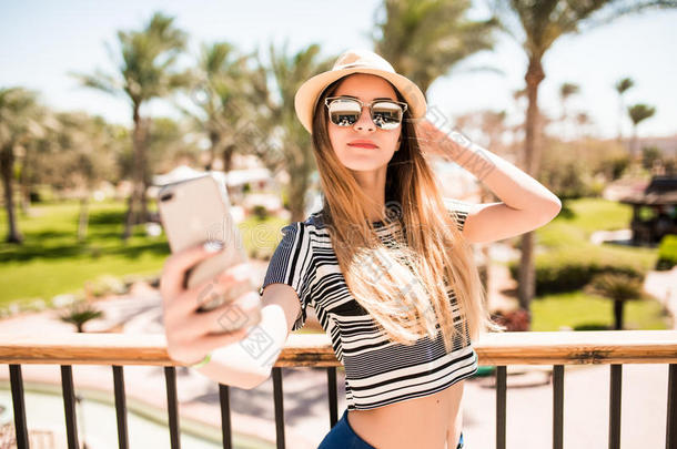 开朗的年轻女人戴着帽子和太阳镜，在夏<strong>季</strong>度假胜地的手掌背景上用手机自拍。 <strong>暑假</strong>