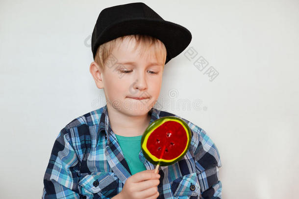 可爱的小男孩穿着时髦的衬衫和黑色的帽子，看着五颜六色的棒棒糖。 可爱的小学生穿着时髦的衣服
