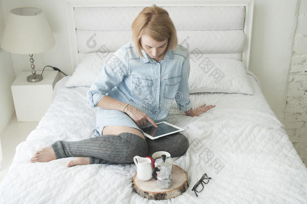 女孩在一张白色的床上喝早咖啡，在高丝袜的平板电脑上工作