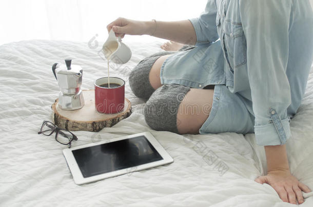 女孩在一张白色床上喝早咖啡，读高丝袜的平板电脑