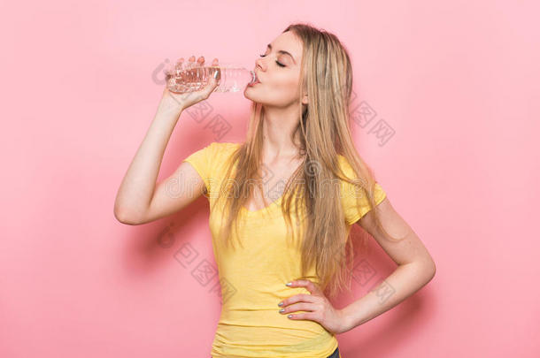 美丽的年轻健身妇女，完美的体型，饮用淡水从塑料瓶站在粉红色的墙壁附近