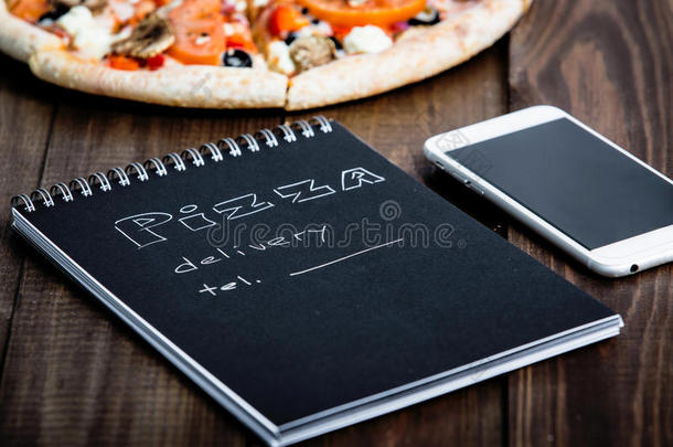 比萨饼的特写，一部手机和一个笔记本，上面写着：比萨饼的送货。 背景木桌。 笔记本黑色