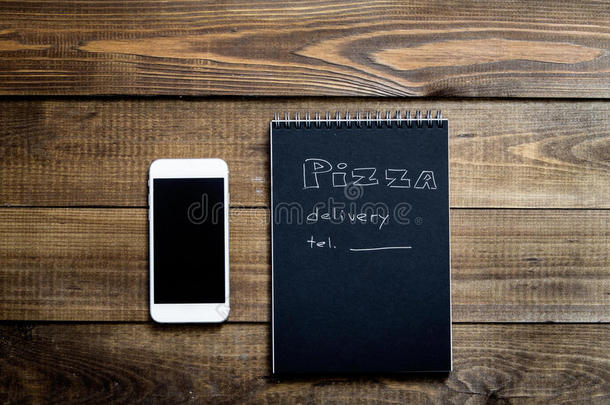 手机和笔记本的特写与文字：比萨饼送货。 背景木桌。 笔记本黑色和白色