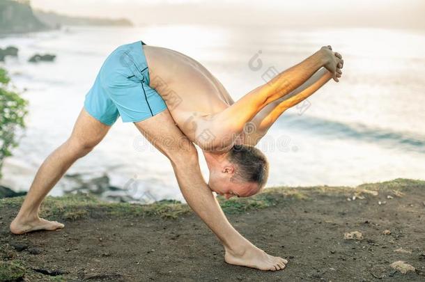 英俊的男人做瑜伽在悬崖和蓝色海背景