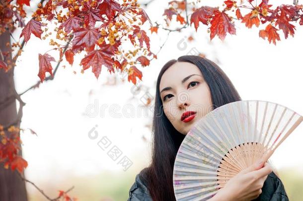 美丽的年幼的亚洲人女人和扇子向背景关于红色的枫树