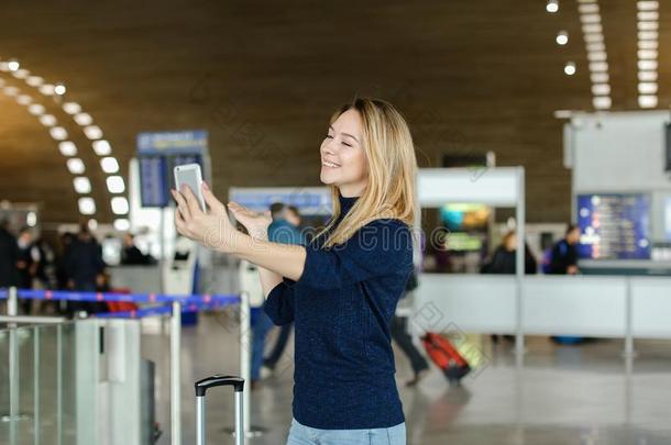 女孩制造自拍照在旁边智能手机在机场过道和旅行小皮包.