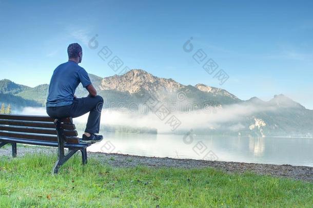 男人坐向木制的长凳在海岸关于湖发出吼叫声蓝色山