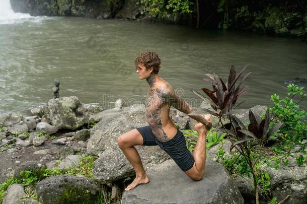 刺青男人开业的瑜伽向岩石和河和绿色的植物