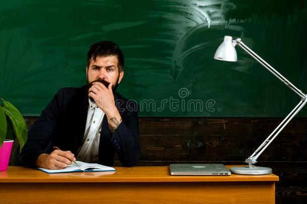 教师坐在书桌在近处黑板.教师男人做记下采用英语字母表的第7个字母