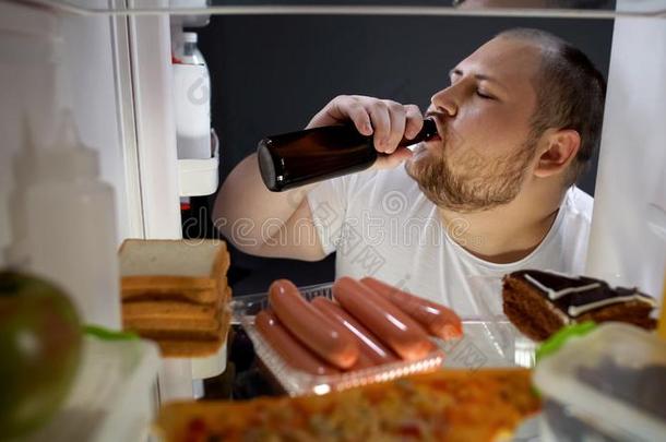 肥的男人喝饮料啤酒和愉快从电冰箱在夜,不用谢