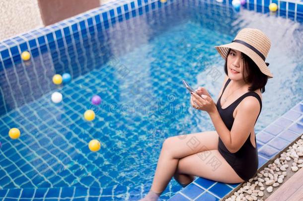美丽的亚洲人女人使用可移动的电话在游泳水池,幸福的