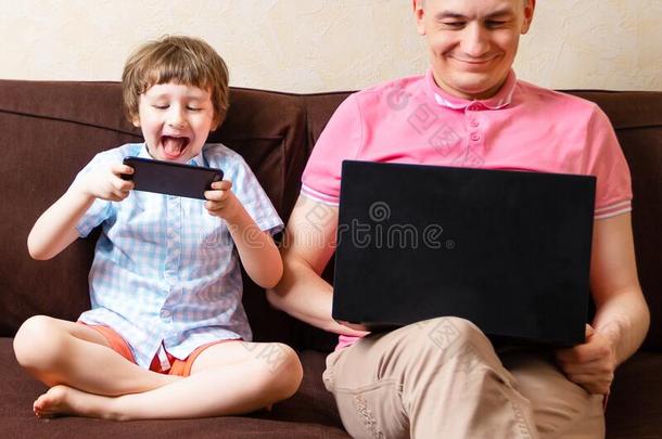 幸福的年幼的父亲坐向长沙发椅使用便携式电脑轻松和<strong>小孩</strong>s向