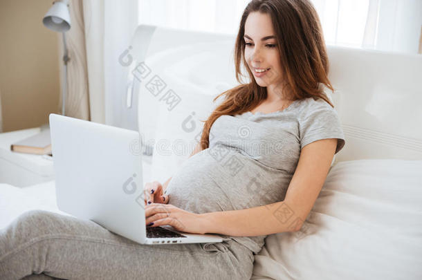 欢乐的怀孕的年幼的女人说谎和使用便携式电脑向床