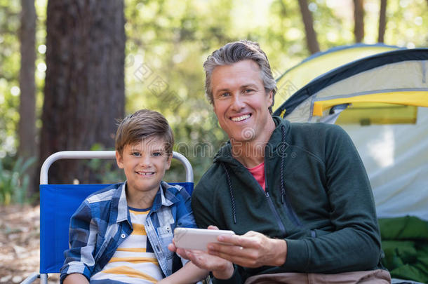 微笑的父亲和儿子使用可移动的电话在旁边帐篷采用森林