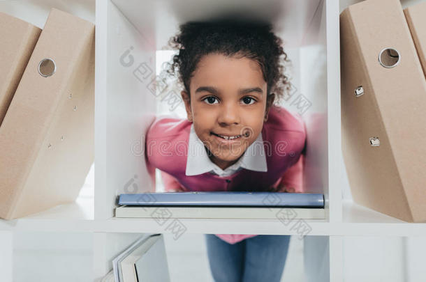 <strong>小</strong>的女孩微笑的在照相机通过<strong>书架</strong>采用办公室