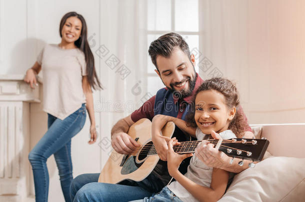 供多种族用的父亲和女儿演奏吉他向沙发在家