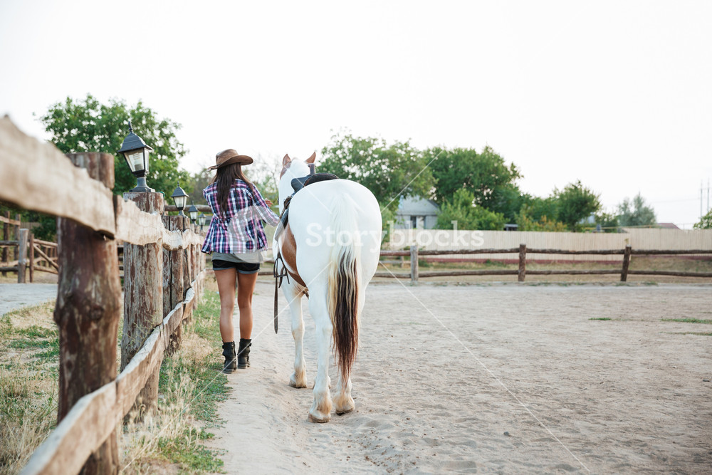 年轻女牛仔在农场中牵着白马一起散步的背影